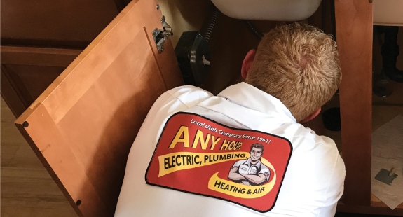 A person wearing a white shirt with the logo Any Hour Electric, Plumbing, Heating & Air repairs a sink under a wooden cabinet. The tone is professional and focused.