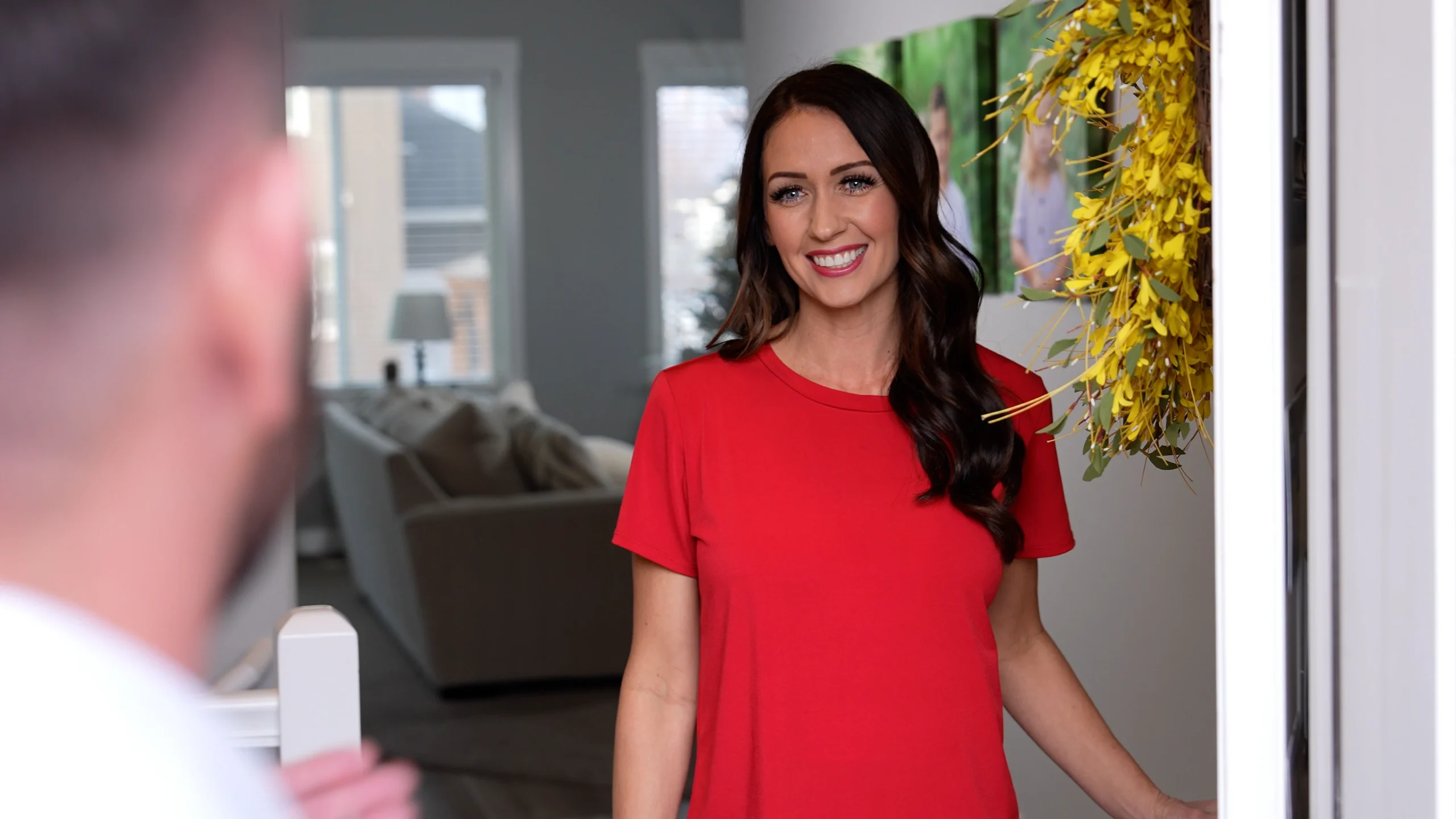 A smiling woman in a red shirt stands welcomingly at the open door of a cozy home, greeting a technician. Yellow flowers decorate the entrance, conveying warmth and hospitality.