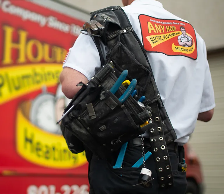 A plumber in a branded AnyHour Services uniform carries a tool bag filled with pliers and other tools, standing beside a colorful company van. Professional and ready.