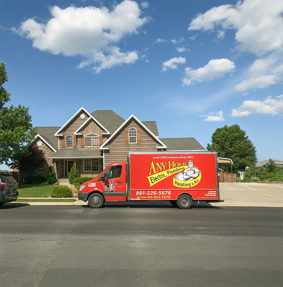 Red service truck labeled Any Hour Electric, Plumbing, Heating & Air parked in front of a large suburban house under a clear blue sky. Peaceful scene.
