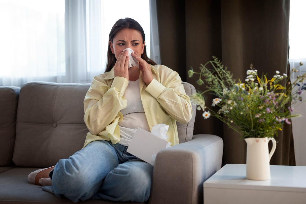 A woman sneezes while sitting on a couch in her living room. There is a vase of wildflowers on the table next to the couch. Spring allergies prevented by IAQ and HVAC.